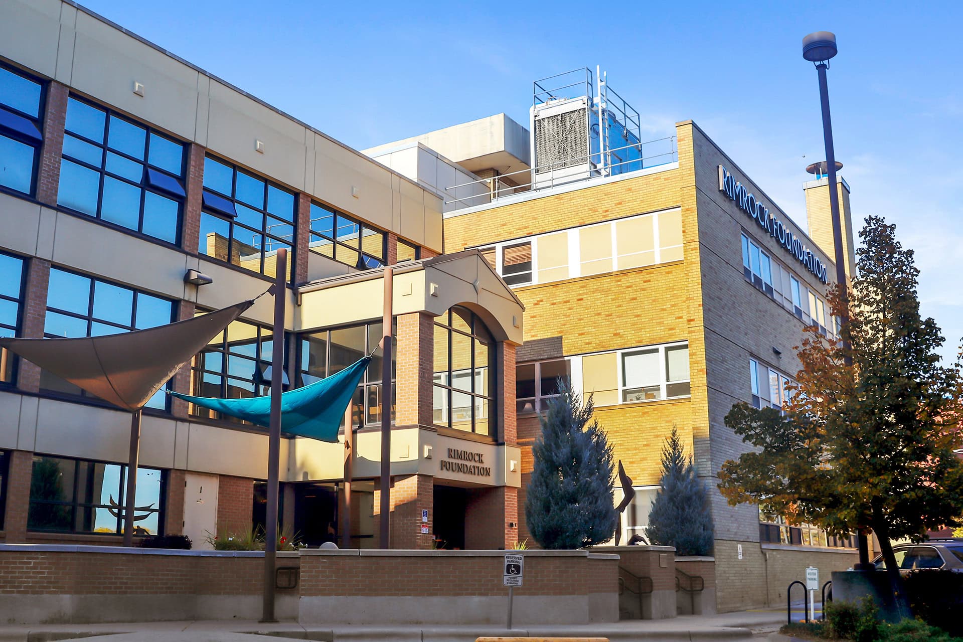 Modern multi-story building of Rimrock Foundation, a leading addiction treatment center in Montana, featuring large glass windows, outdoor shade sails, and well-maintained landscaping on a sunny day.